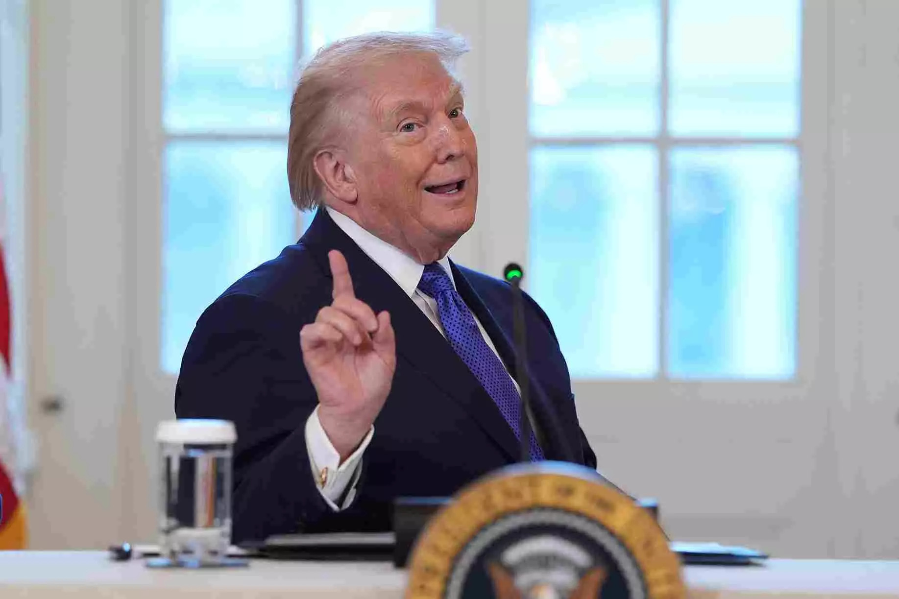 Donald Trump speaking at a meeting table, raising one finger while addressing an audience indoors